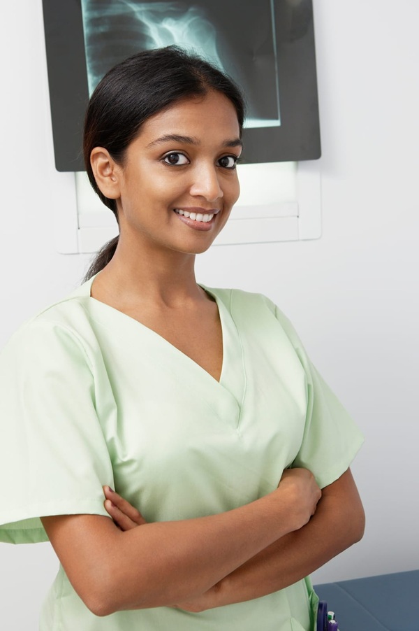 A woman in light green scrubs stands with arms crossed, smiling, in a medical setting with an X-ray image visible in the background.