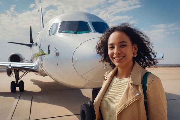 A woman stands smiling in front of a parked airplane on an airport tarmac under a partly cloudy sky, embodying the adventure of learning how to become a travel nurse.