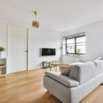 Minimalist living room with a gray sofa, wall-mounted TV, two windows with blinds, light wood flooring, and a small white shelving unit with decor.
